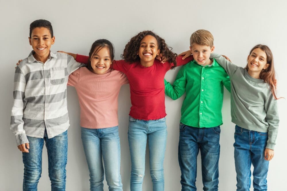 Cheerful Multiethnic Children Hugging Posing Smiling To Camera Indoors. Preteen Boys And Girls Standing Together Over Gray Wall. Childhood And Friendship